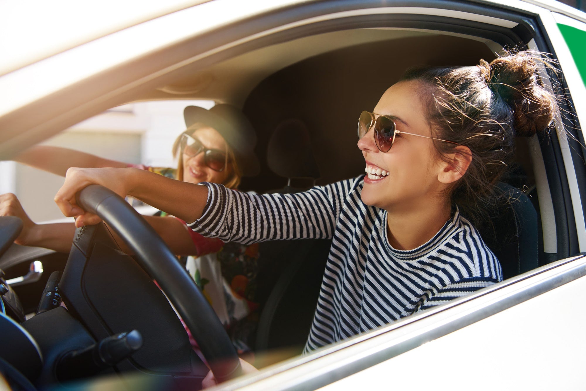 Kaksi naista istuvat iloisina autossa aurinkolasit päässä